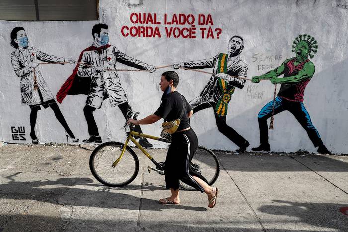 Una mujer pasa al frente de un graffiti que representa al presidente brasileño Jair Bolsonaro y una figura que representa el nuevo coronavirus tirando de una cuerda contra los trabajadores de la salud con un mensaje que dice: "¿De qué lado de la cuerda estás?", en São Paulo (Brasil). EFE/ Sebastião Moreira.