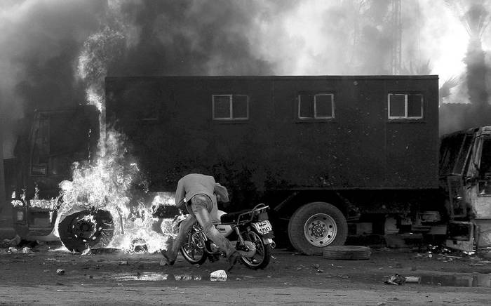 Un hombre con su moto pasa junto a un coche de la Policía en llamas durante las manifestaciones, el viernes, en Suez, a unos 100 kilómetros de El Cairo.
 · Foto: EFE, s/d de autor
