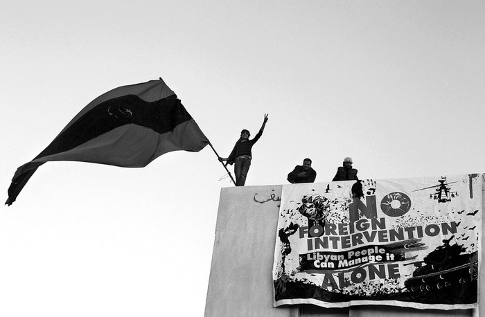 Un joven hace ondear una bandera anterior a la era Gadafi durante una concentración de opositores al régimen, convocada en Bengasi,
junto a él cuelga un cartel en el que puede leerse en inglés: “No a la intervención extranjera. El pueblo libio puede manejarse solo”.
 · Foto: Efe, Tiago Petinga